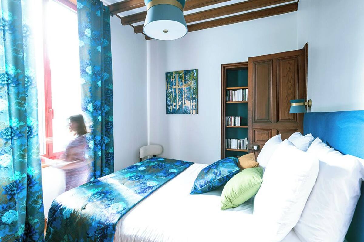 Un dormitorio con cama, estantería, un cuadro y ventana con cortinas.