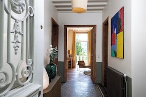 A hallway with a wooden door, a chair, a vase with flowers, and a colorful painting on the wall.