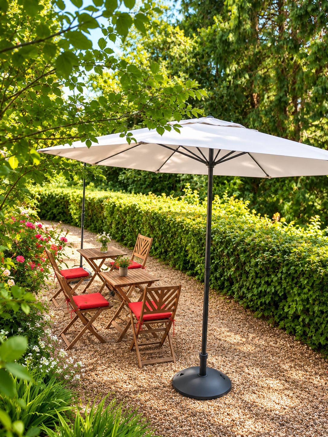 An outdoor seating area with a table and chairs under a large umbrella, surrounded by greenery and a hedge.