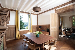 A spacious room with a large wooden dining table, chairs, a vase with flowers, and a view of the outdoors through a window.