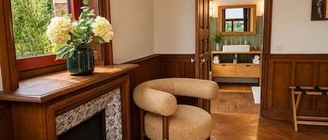 A room with a wooden floor, a fireplace, a wicker chair, a small table with a plant, and a view of a bathroom with a mirror and sink.