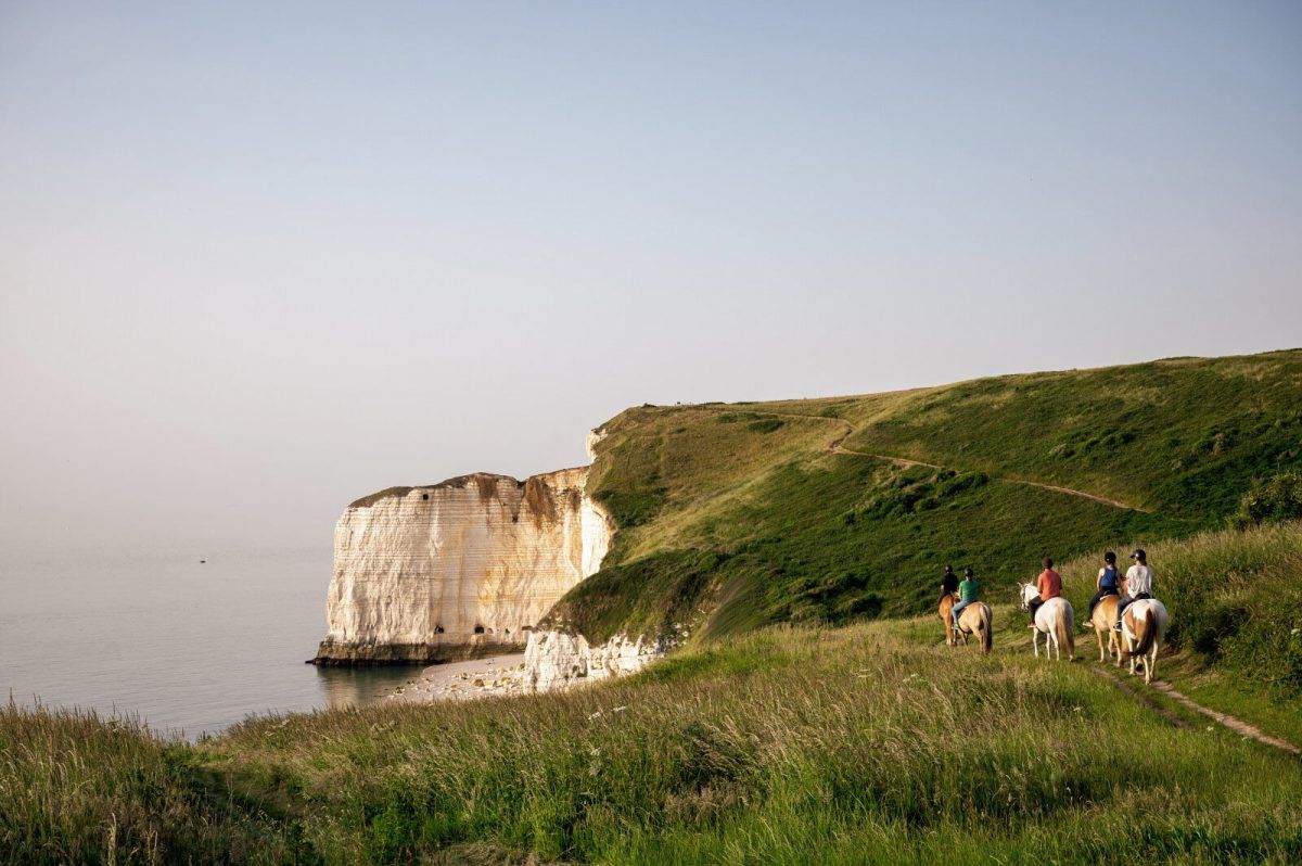 Un paysage côtier caractérisé par une falaise blanche imposante et un sentier où l’on peut voir des gens à cheval.