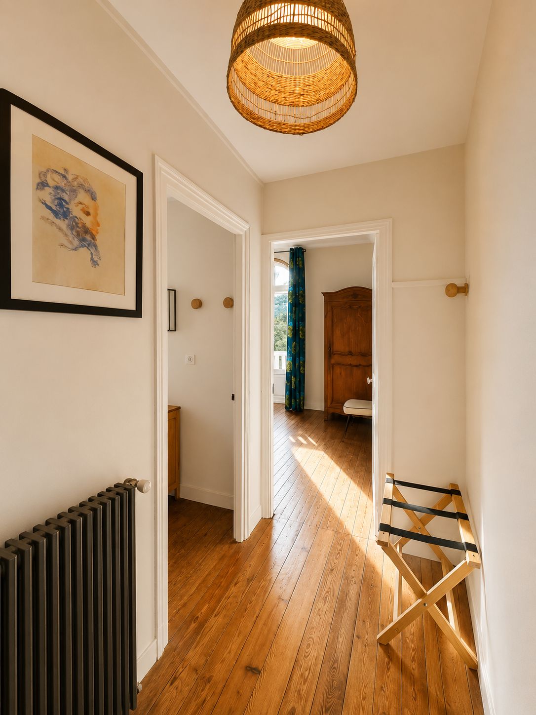 A hallway with a wooden floor, a radiator, a framed artwork, a wicker ceiling light, and a door leading to a bathroom with a wooden cabinet and a shower curtain.