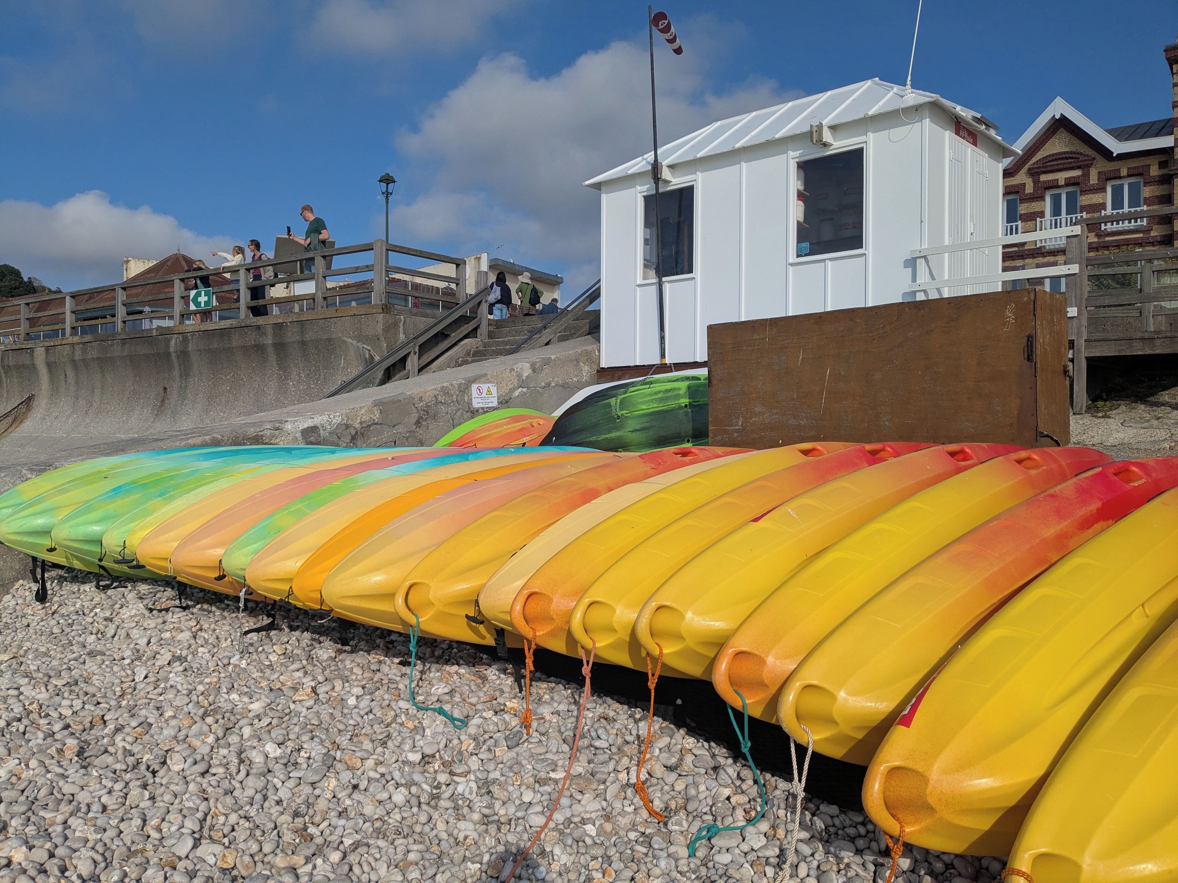 A row of colorful kayaks lined up on a pebbled beach, a lifeguard tower, and a wooden structure.