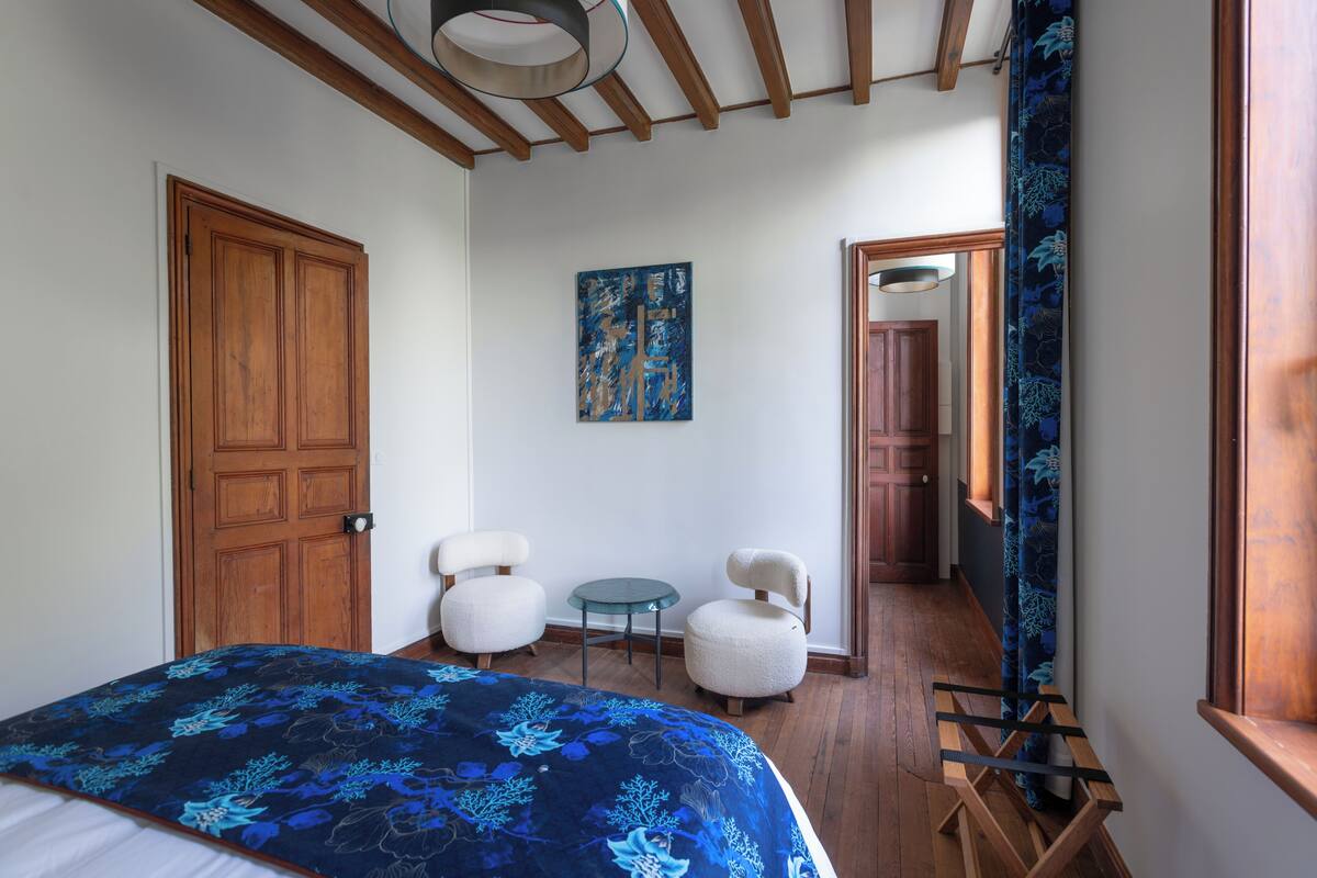 Un dormitorio con puerta de madera, una cama con colcha estampada de flores azules, dos sillas blancas, una mesita redonda y un cuadro en la pared.