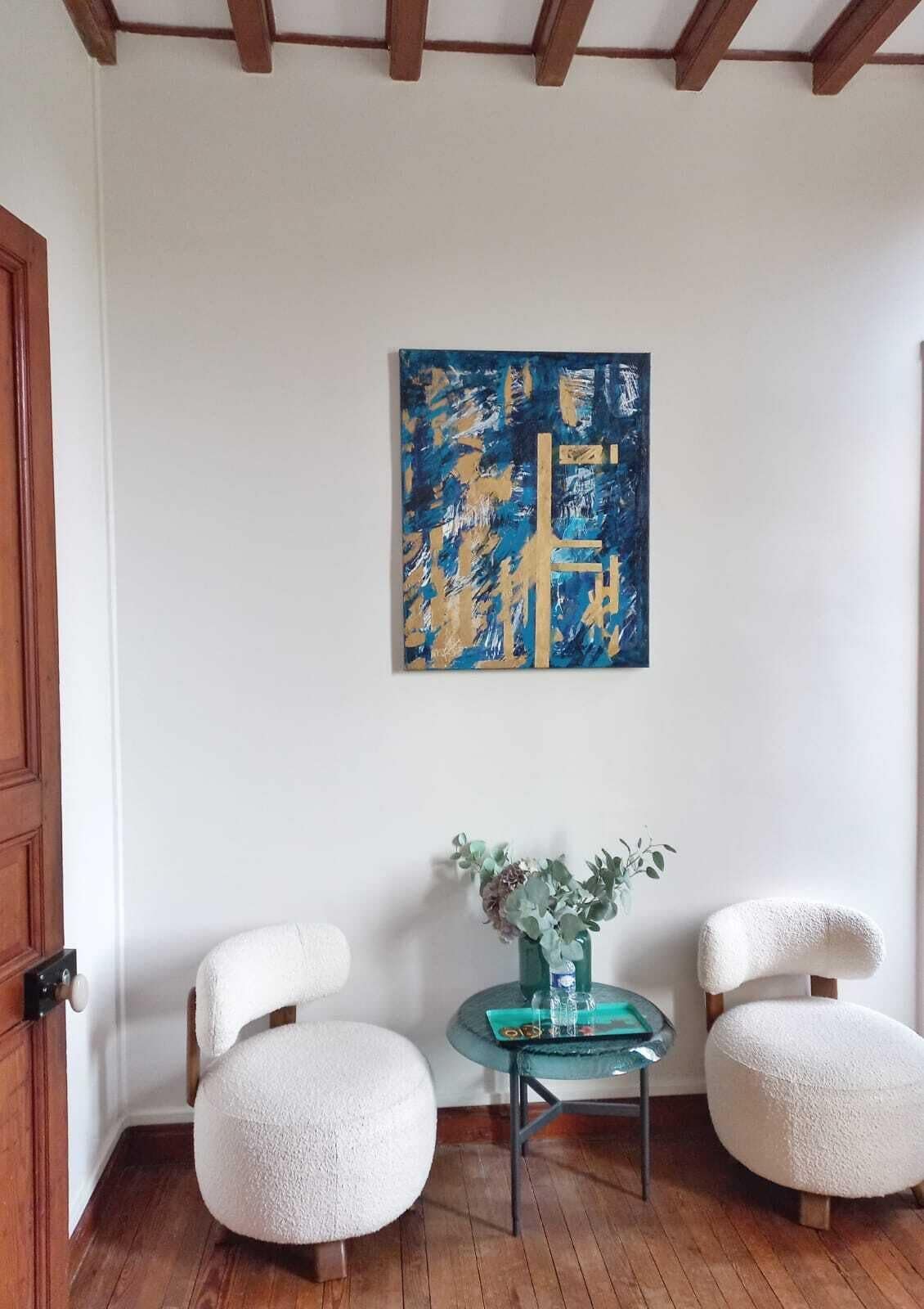 A room with two white chairs, a small table with a vase, and a blue abstract painting on the wall.