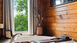 Interior de uma cabana de madeira com banheira, janela com cortinas, um vaso com galhos e um quadro na parede.