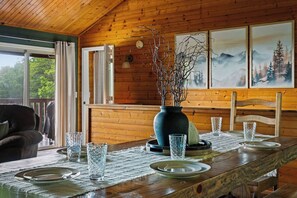 Ein Interieur einer Holzhütte mit einem Esstisch, der für vier Personen gedeckt ist, einer Vase mit Ästen und gerahmten Bildern an der Wand.