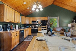 A kitchen with wooden cabinets, stainless steel appliances, and a dining area with a table set for four. - Epic Views•Hot Tub•Game Room•Private•Near PF, Gatlinburg & GSMNP (Sevierville)