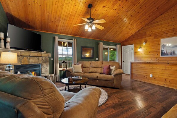 A cozy living room with a stone fireplace, leather sofas, a ceiling fan, and a large TV. - Epic Views•Hot Tub•Game Room•Private•Near PF, Gatlinburg & GSMNP (Sevierville)