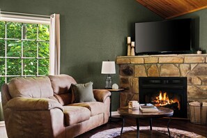 A living room with a stone fireplace, a sofa, a television, and a table with books and a candle. - Epic Views•Hot Tub•Game Room•Private•Near PF, Gatlinburg & GSMNP (Sevierville)
