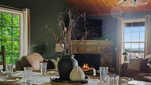 Uma sala de jantar com lareira, um vaso com galhos e uma mesa posta com pratos e copos.