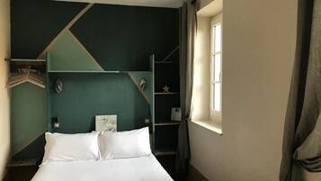 Una cama con sábanas blancas, una pared de color aguamarina y una ventana con cortinas.