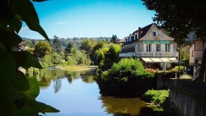 A hotel by a calm river with a view of the surrounding greenery and hills.