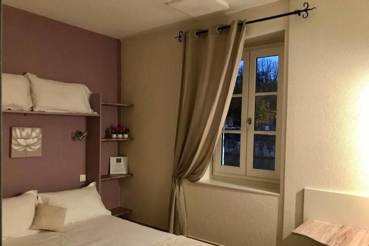 A bedroom with a bed, a nightstand, a window with curtains, and a wall-mounted shelf with decorative items.