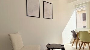 Ett rum med en vit stol, ett litet bord, två inramade konstverk på väggen och ett fönster med utsikt över byggnader.
