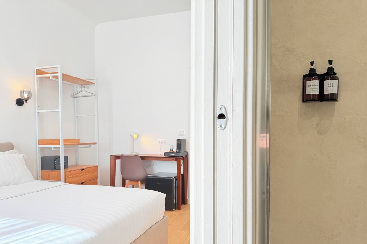 A hotel room with a bed, a desk with a chair, a wardrobe, and a wall-mounted shelf with two bottles.