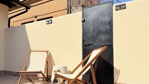 En uteplats med två trästolar och ett litet bord, en svart grind och en beige vägg.