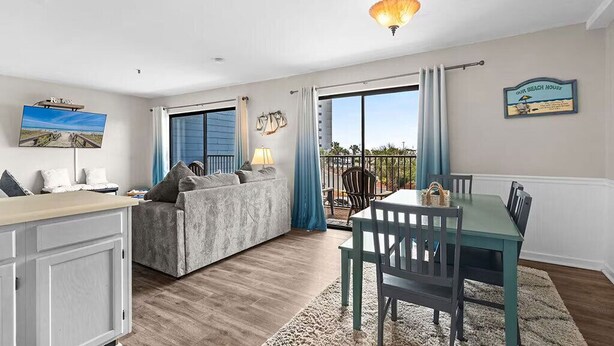 A living room with a sofa, a dining table with chairs, a TV mounted on the wall, and a balcony with a view.