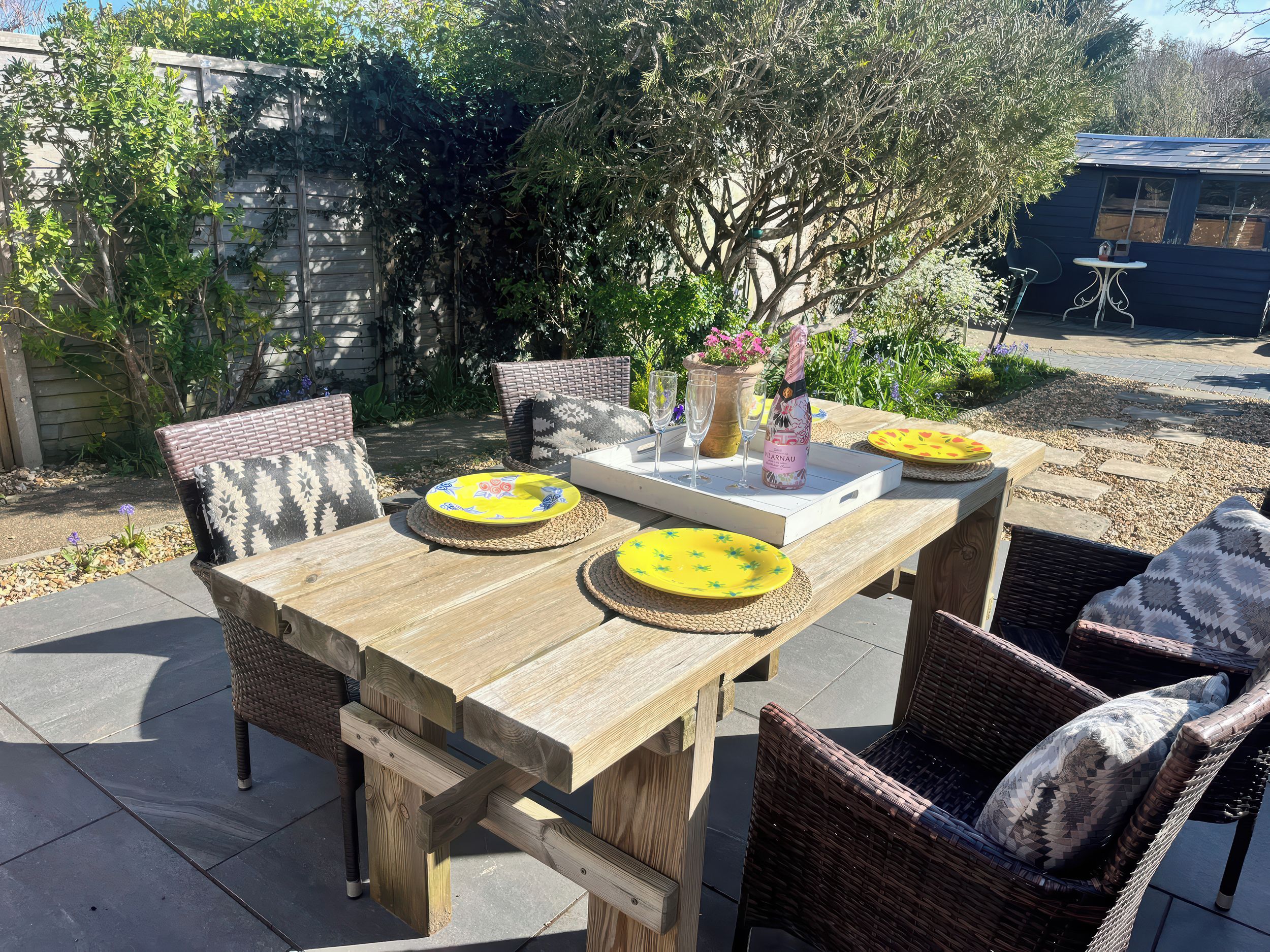 A wooden patio table with chairs, a bottle of champagne, and plates.