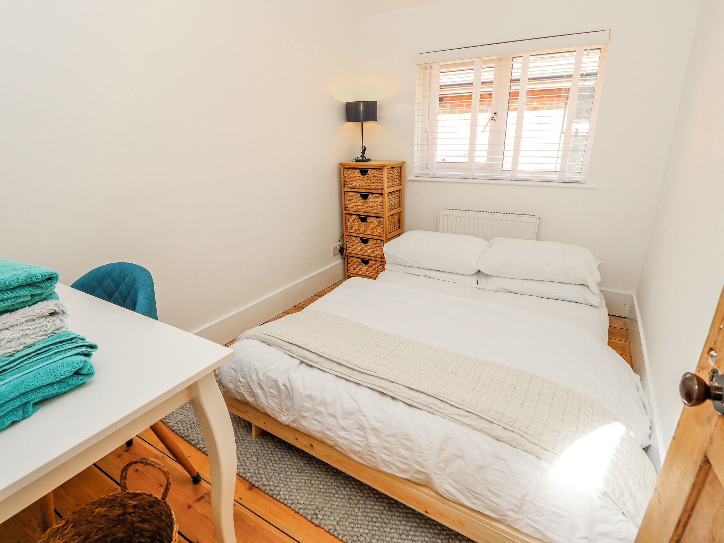 Een slaapkamer met een bed, nachtkastje, lamp, raam met zonwering en een kleine houten dressoir.
