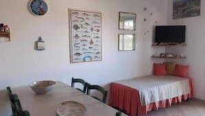 Una habitación con una mesa de comedor, sillas, una cama con colcha roja, un televisor de pared y un cartel de un pez.