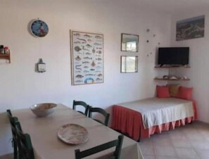 Una habitación con una mesa de comedor, sillas, una cama con colcha roja, un televisor de pared y un cartel de un pez.