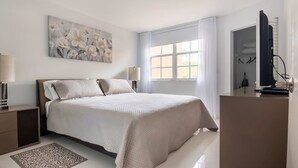 Ein Schlafzimmer mit Bett, Nachttischen, Schreibtisch, Stuhl, einem Fenster mit Vorhängen, einem Bild an der Wand und einer Deckenleuchte.