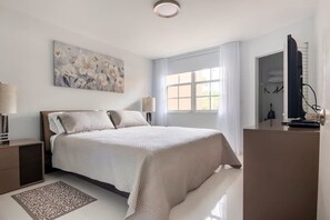 Ein Schlafzimmer mit Bett, Nachttischen, Schreibtisch, Stuhl, einem Fenster mit Vorhängen, einem Bild an der Wand und einer Deckenleuchte.