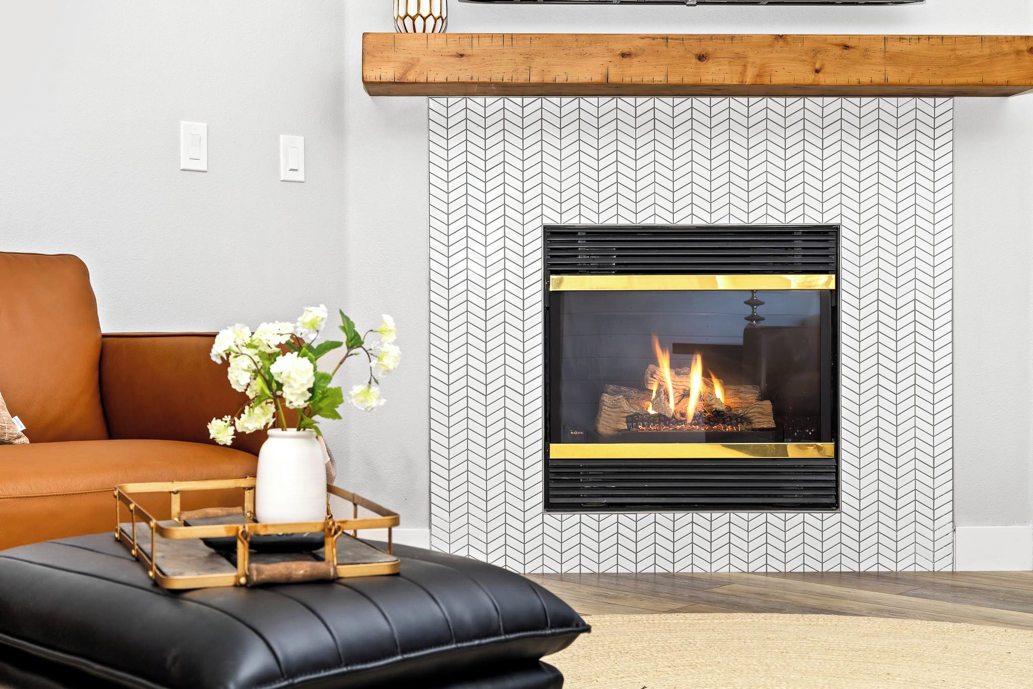 A modern living room with a fireplace, a brown leather sofa, a black leather ottoman, and a white vase with flowers.