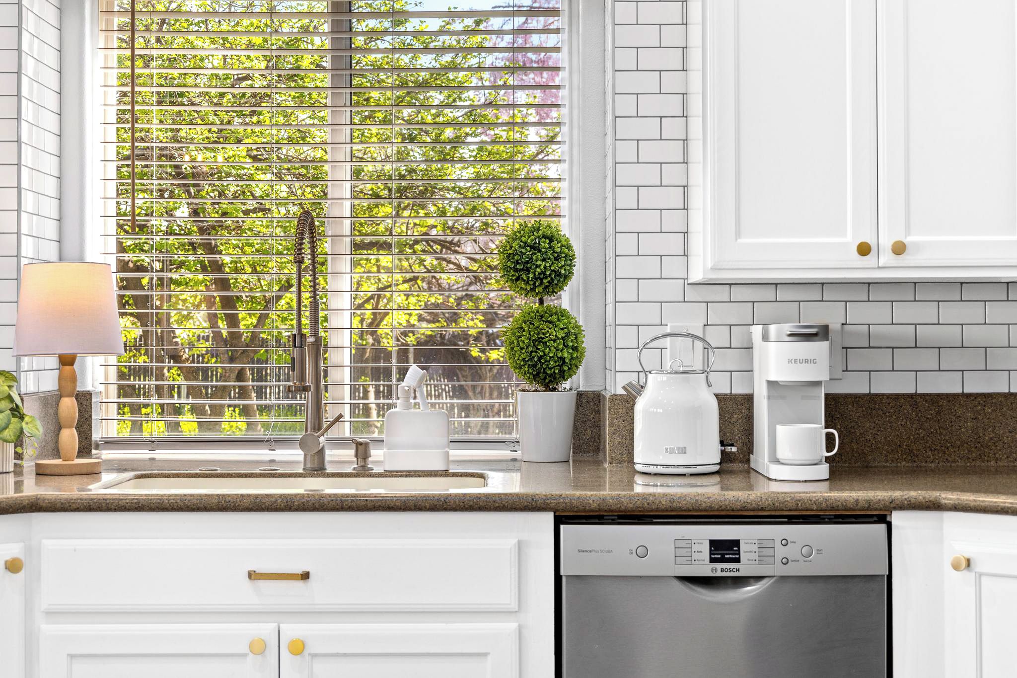 A kitchen with white cabinets, a Bosch dishwasher, a Keurig coffee maker, a potted plant, and a window with blinds.