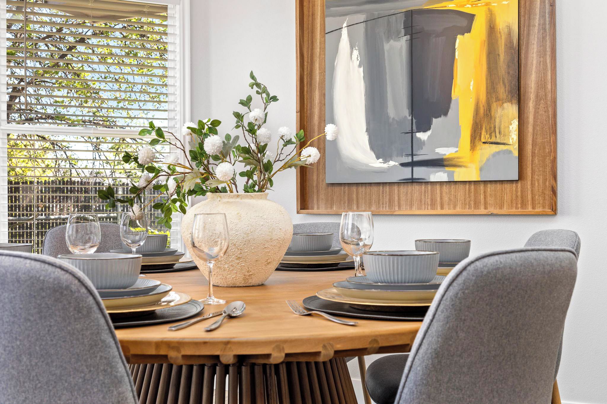 A dining area with a wooden table set for four, a vase with white flowers, and a modern abstract painting on the wall.
