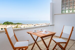 Een balkon met een tafel en stoelen, uitzicht op zee, en een raam met een rasterpatroon.