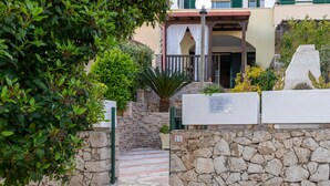 Un muro de piedra con un cartel, un acceso pavimentado y un edificio con un balcón.