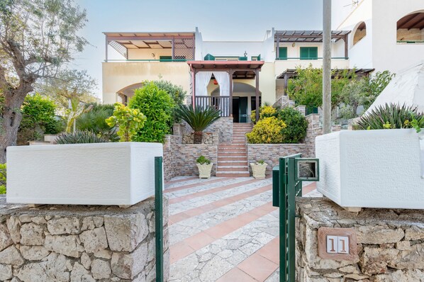 A modern building with a stone pathway leading to a gated entrance, surrounded by greenery and a clear sky.