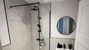 A modern bathroom with a glass-enclosed shower, a round mirror, a sink with a small plant, and a teal rug. - Modern 1BR Ground Floor Flat | City Centre Near High Street (Southend-on-Sea)
