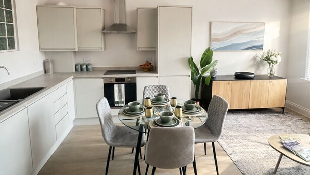 A modern kitchen with white cabinetry, a dining area with a glass table and grey chairs, and a view of the sea through a window. - Modern 1BR Ground Floor Flat | City Centre Near High Street (Southend-on-Sea)
