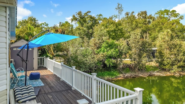 A deck with a blue umbrella, white railing, and a view of a lake surrounded by trees. - Lakeside Lily Pad (Fort Myers)
