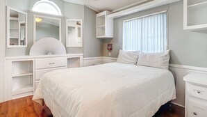 A bedroom with a bed, a vanity with a mirror, a ceiling fan, and a window with curtains. - Lakeside Lily Pad (Fort Myers)