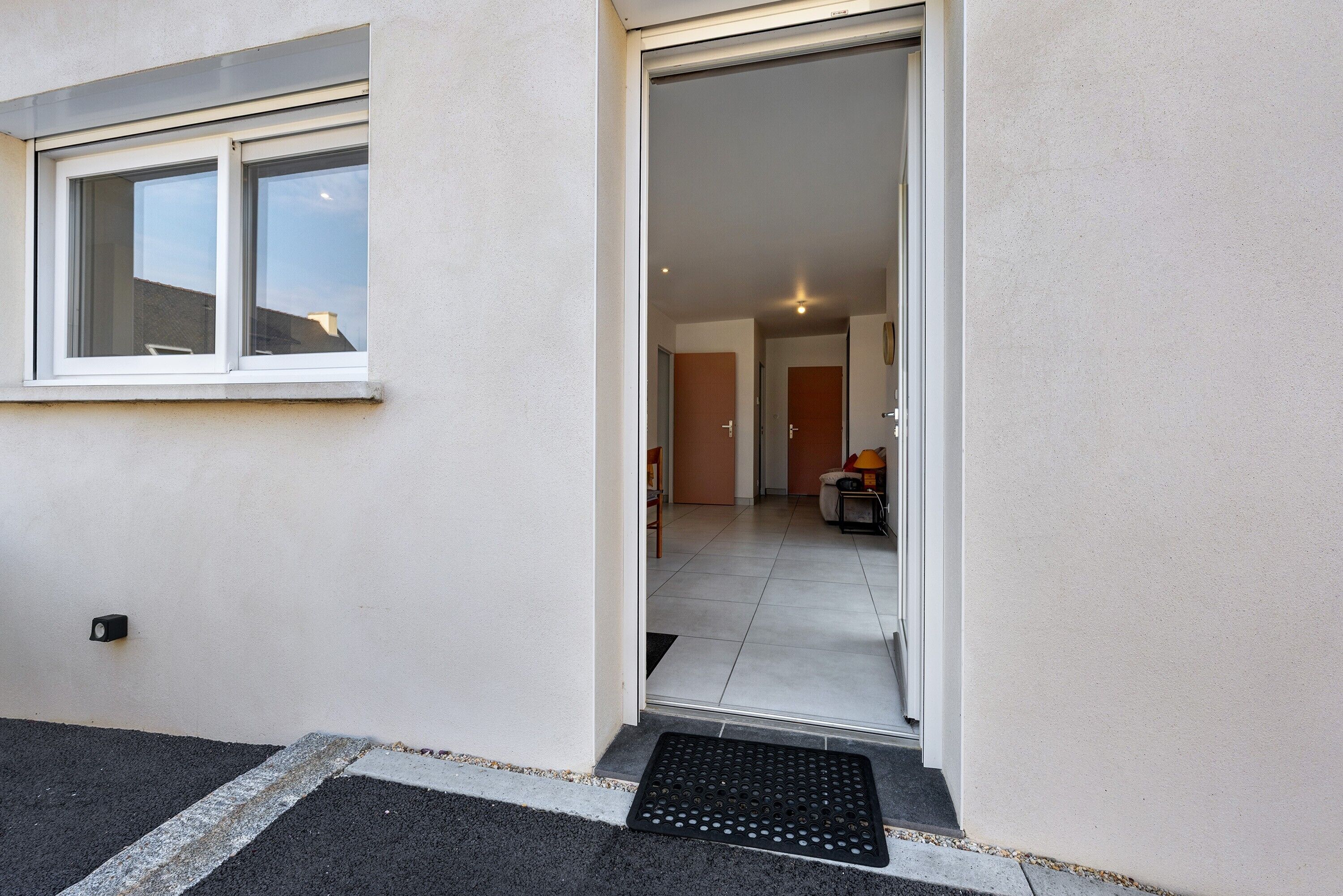 A modern building entrance with a sliding glass door, a small outdoor area with a mat, and a visible interior with a tiled floor and furniture.