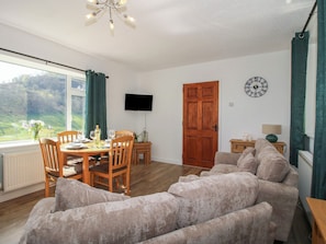 A living room with a sofa, a dining table with chairs, a TV mounted on the wall, and a wooden door. - Spring Bank (Llanfyllin)