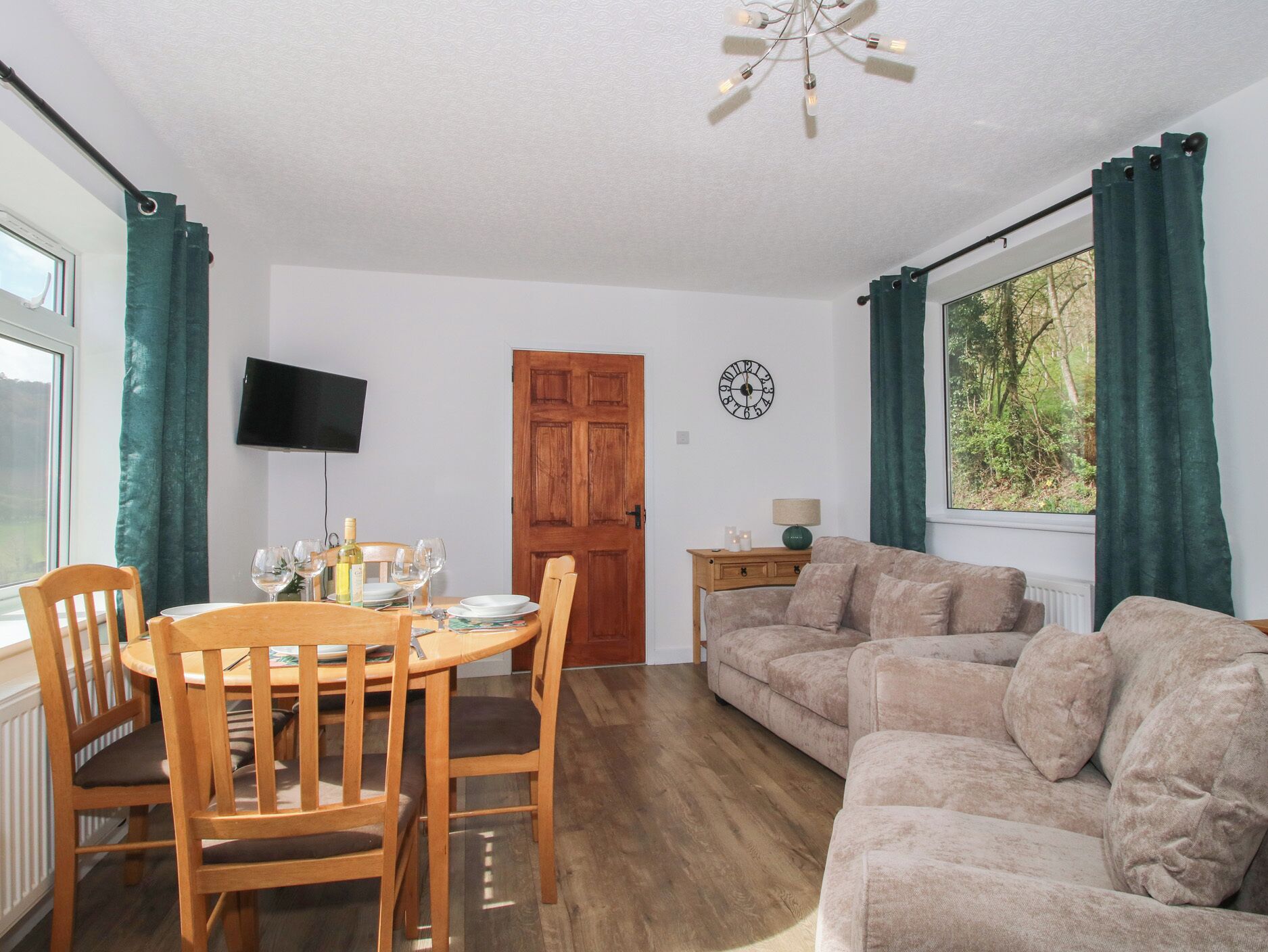 A dining area with a wooden table and chairs, a sofa, a TV mounted on the wall, and a window with curtains.
