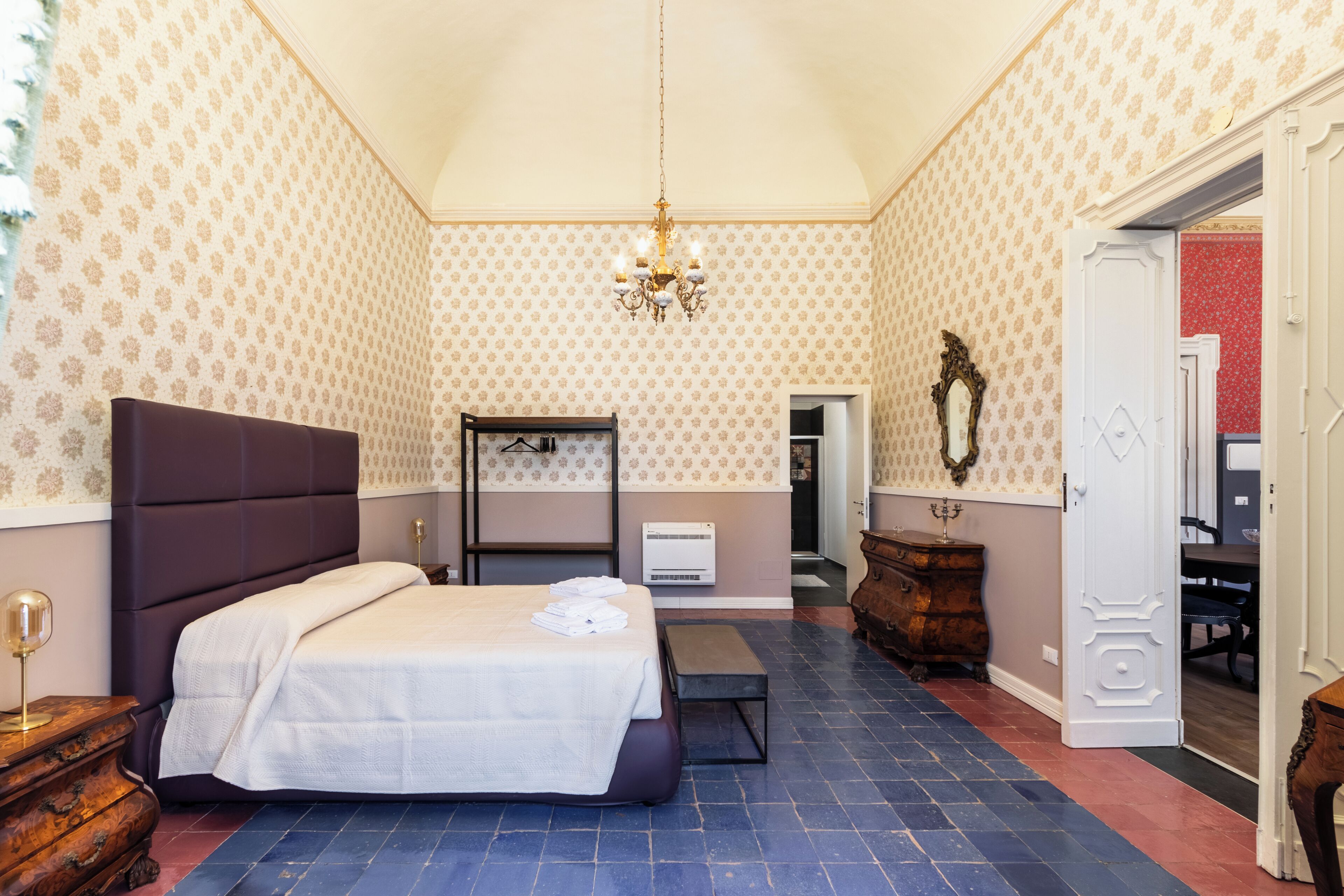 A bedroom with a bed, a bench, a dresser, a mirror, a chandelier, and a door leading to another room.