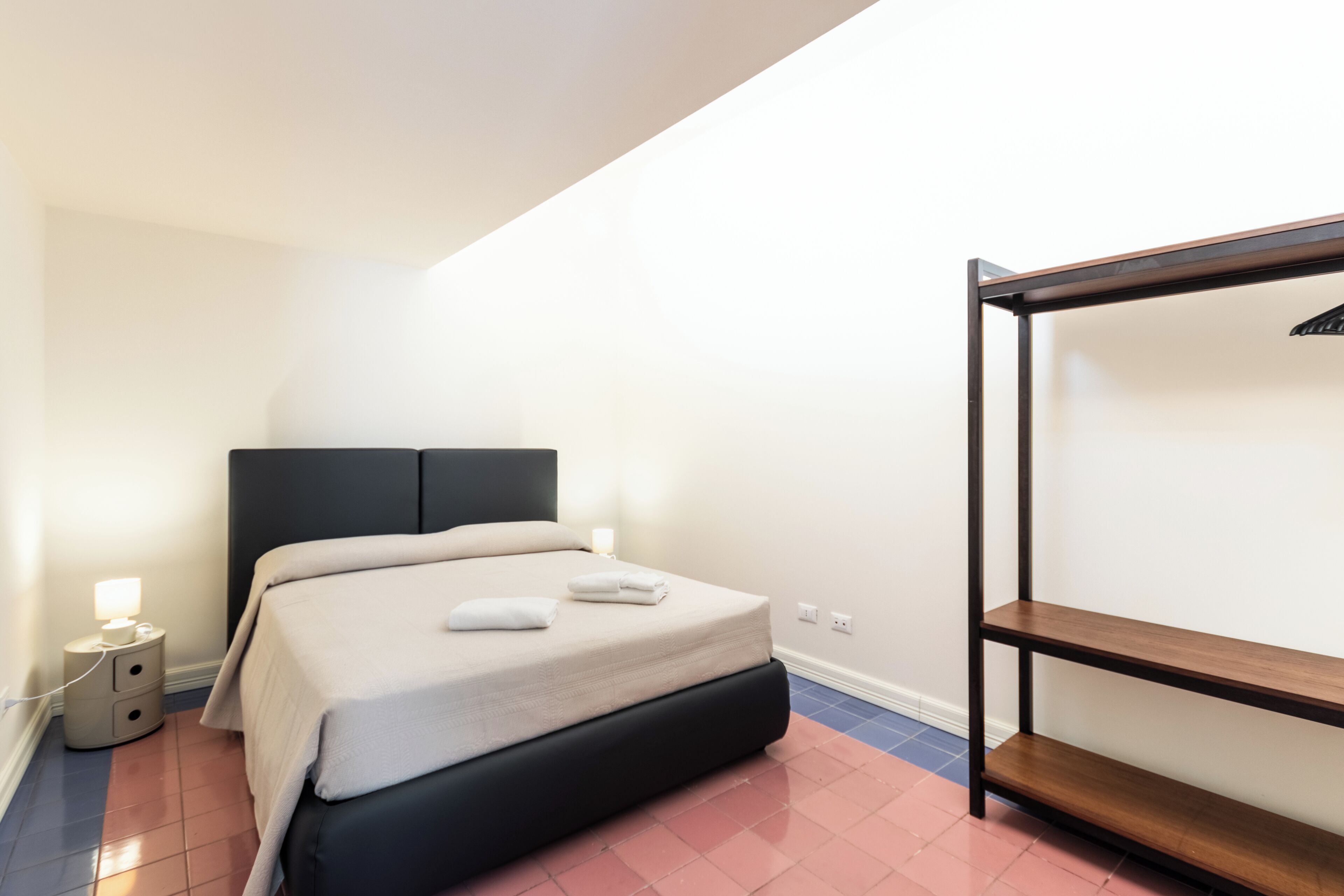 A bedroom with a bed, bedside table, lamp, and a wooden shelf.
