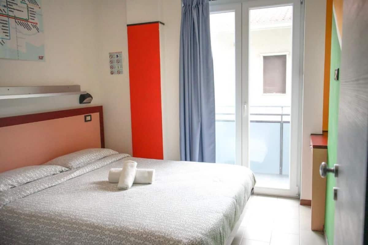 A hotel room with a bed, a red accent wall, a window with curtains, and a view of the outside.