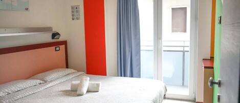 A hotel room with a bed, a red accent wall, a window with curtains, and a view of the outside.