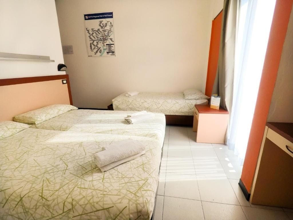 A hotel room with two beds, a small table, a nightstand, a poster on the wall, and a window with curtains.