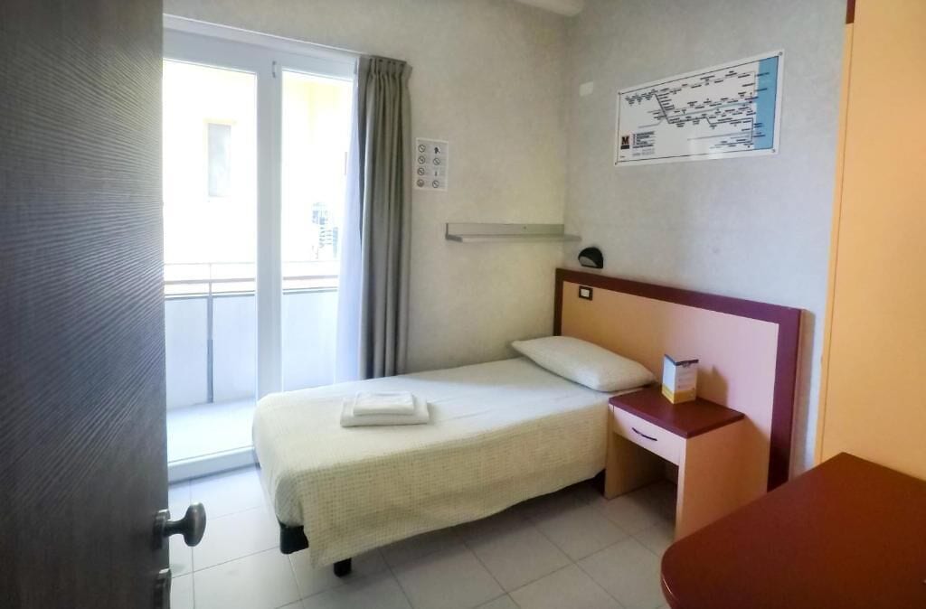 Een hotelkamer met een bed, nachtkastje, een klein tafeltje, een raam met gordijnen en een aan de muur hangende kaart.