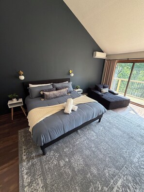A modern bedroom with a dark-colored bed, a small bedside table, a nightstand, and a large window with a view of greenery.