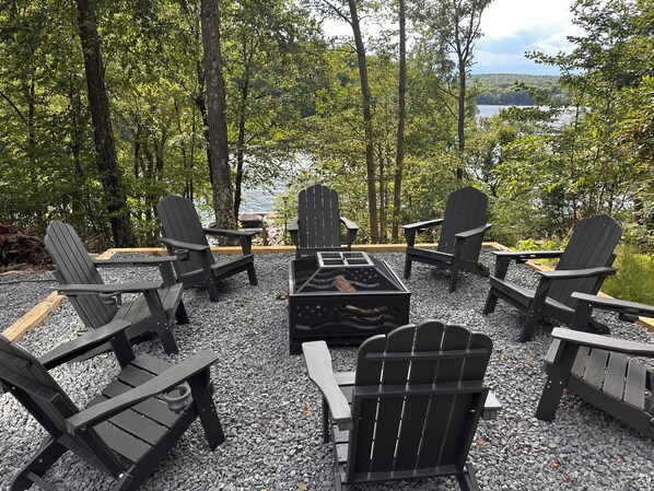 A circular seating arrangement with Adirondack chairs around a central fire table, set on a gravel surface with a lake and forest in the background.